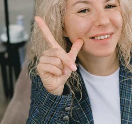 Crop happy adult female entrepreneur with blond hair in formal clothes smiling and showing index finger up sign while looking at camera sitting in armchair in cafe