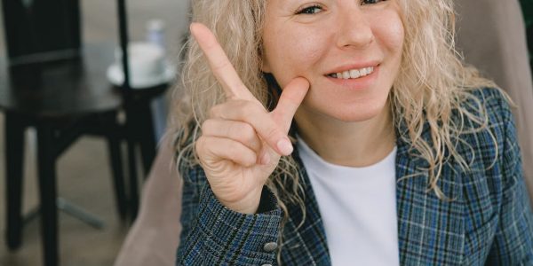 Crop happy adult female entrepreneur with blond hair in formal clothes smiling and showing index finger up sign while looking at camera sitting in armchair in cafe